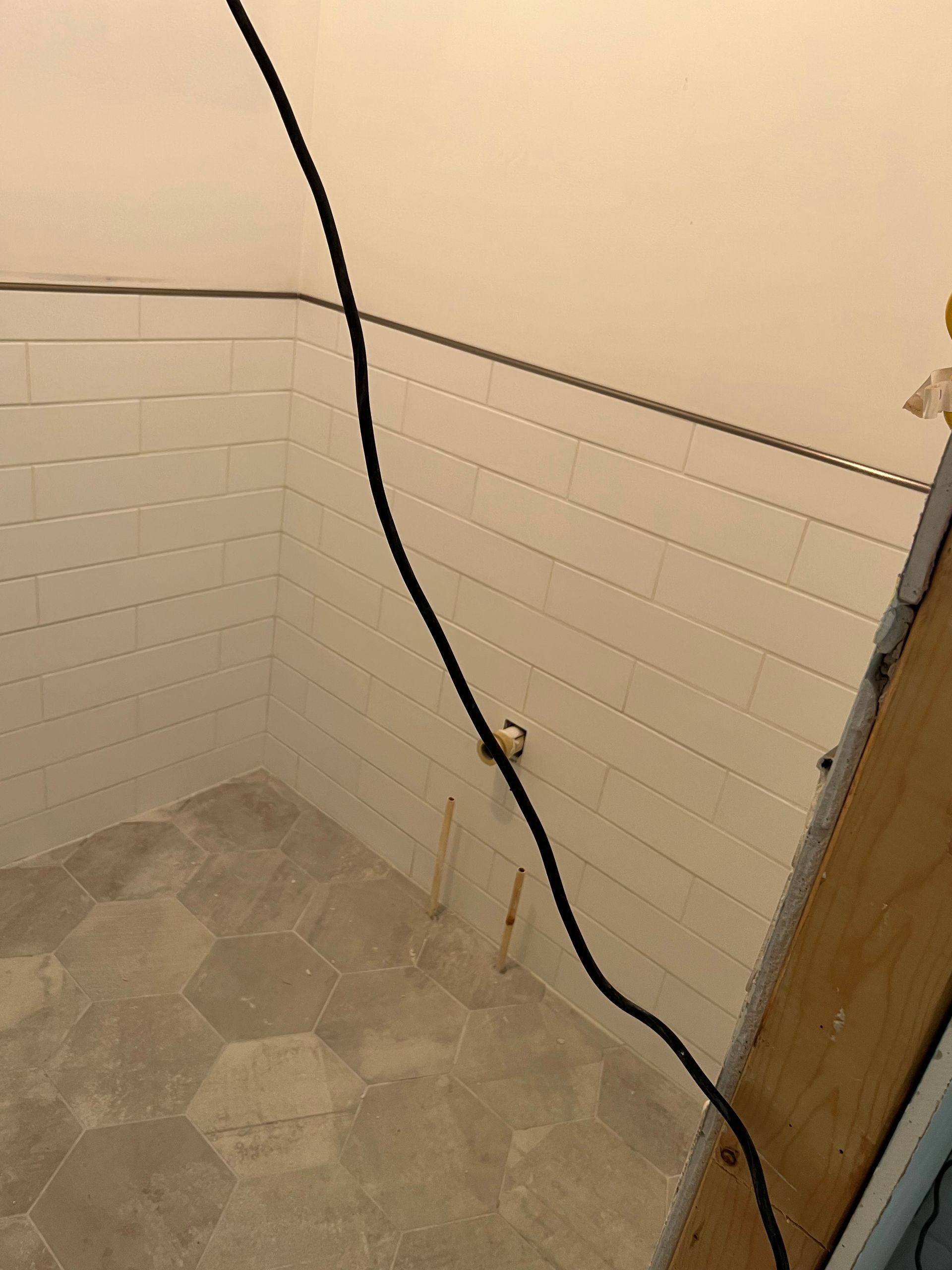 Small tiled shower corner with a black cord hanging down beside a wooden door frame.