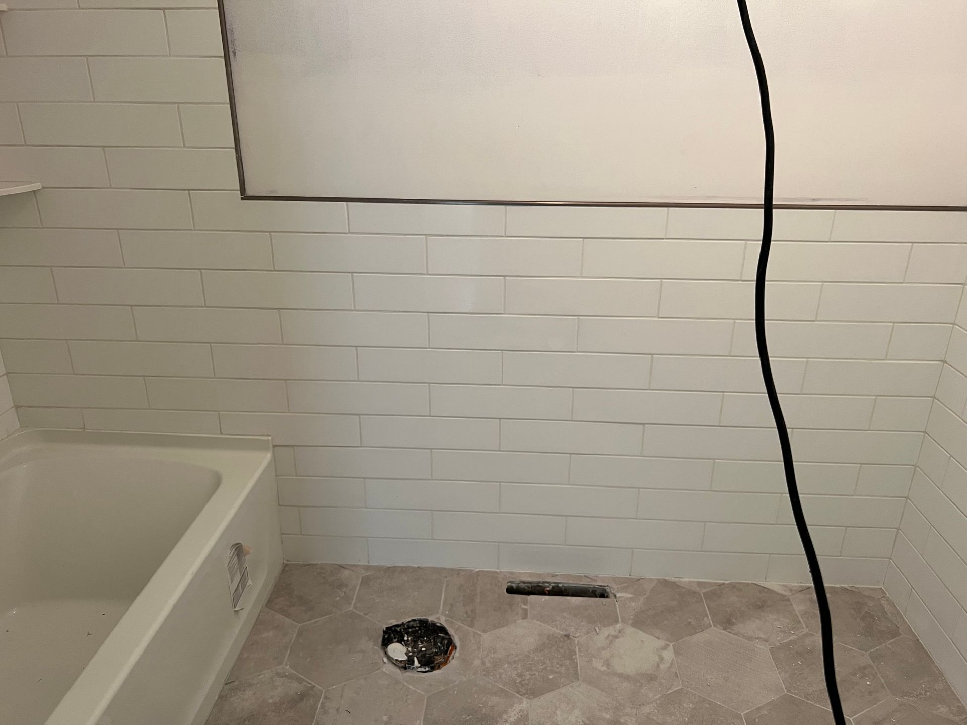 Partially tiled bathroom wall with exposed plumbing, floor drain, and bathtub edge