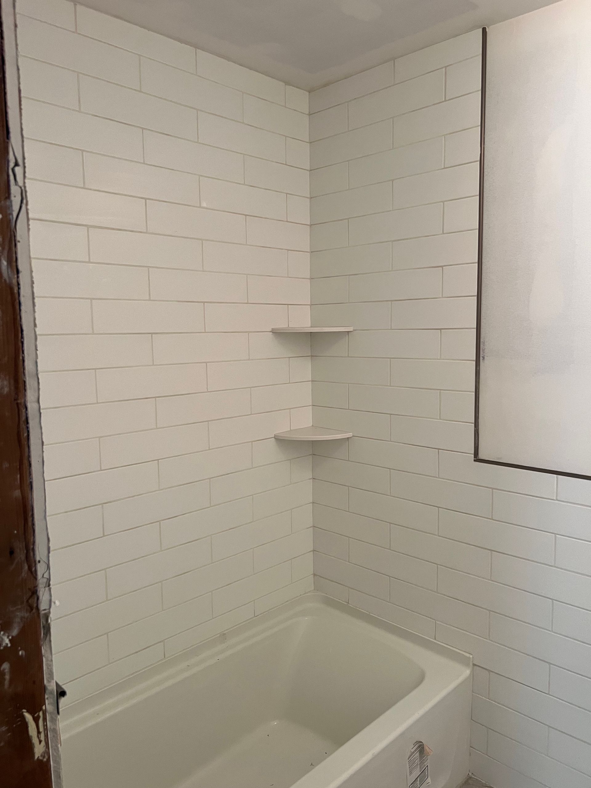 Small white bathroom with a bathtub, tiled walls, and corner shelves near a window.