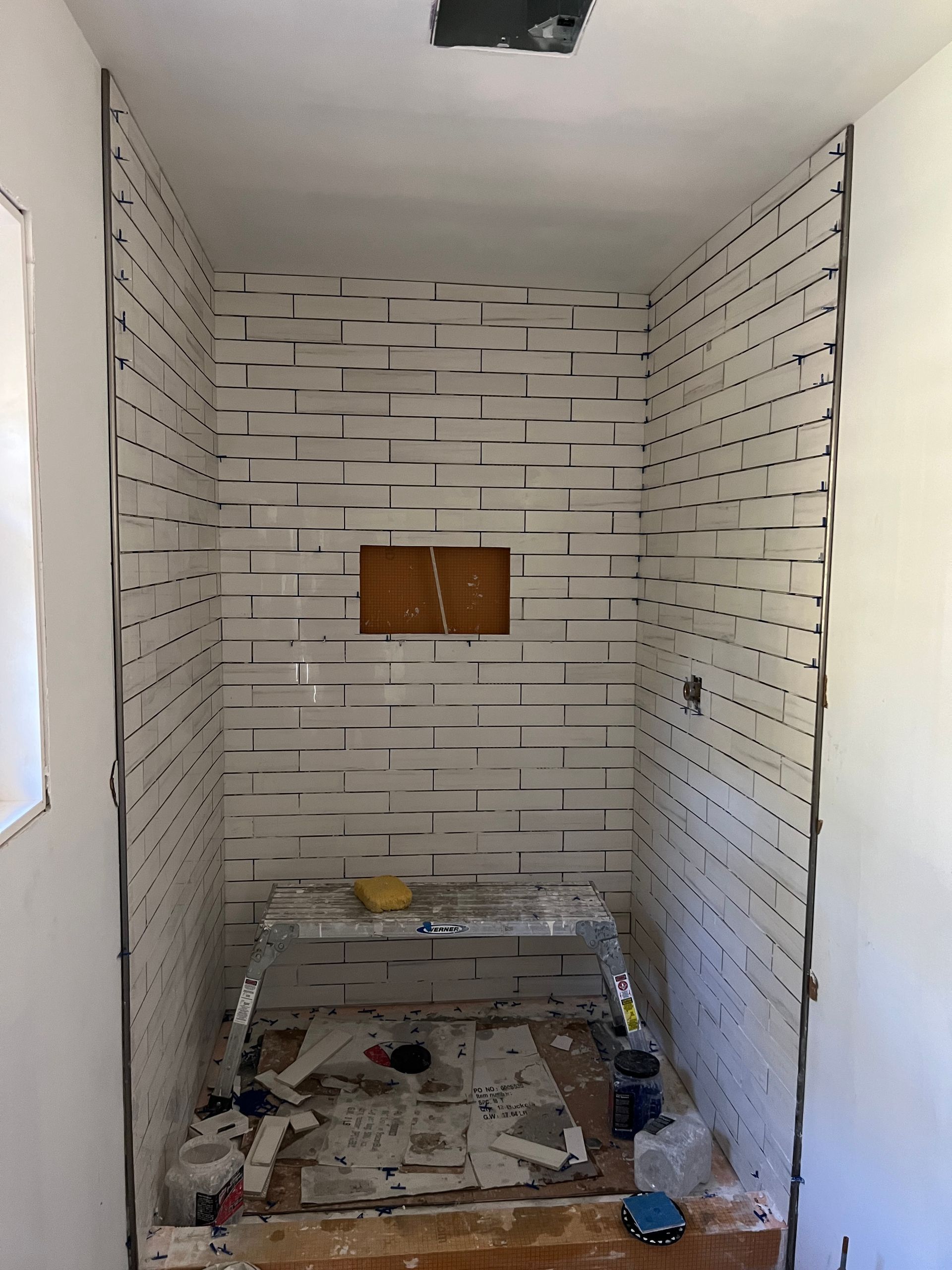 Narrow unfinished room with white tiled walls, exposed floor, and a small wall opening at the far end