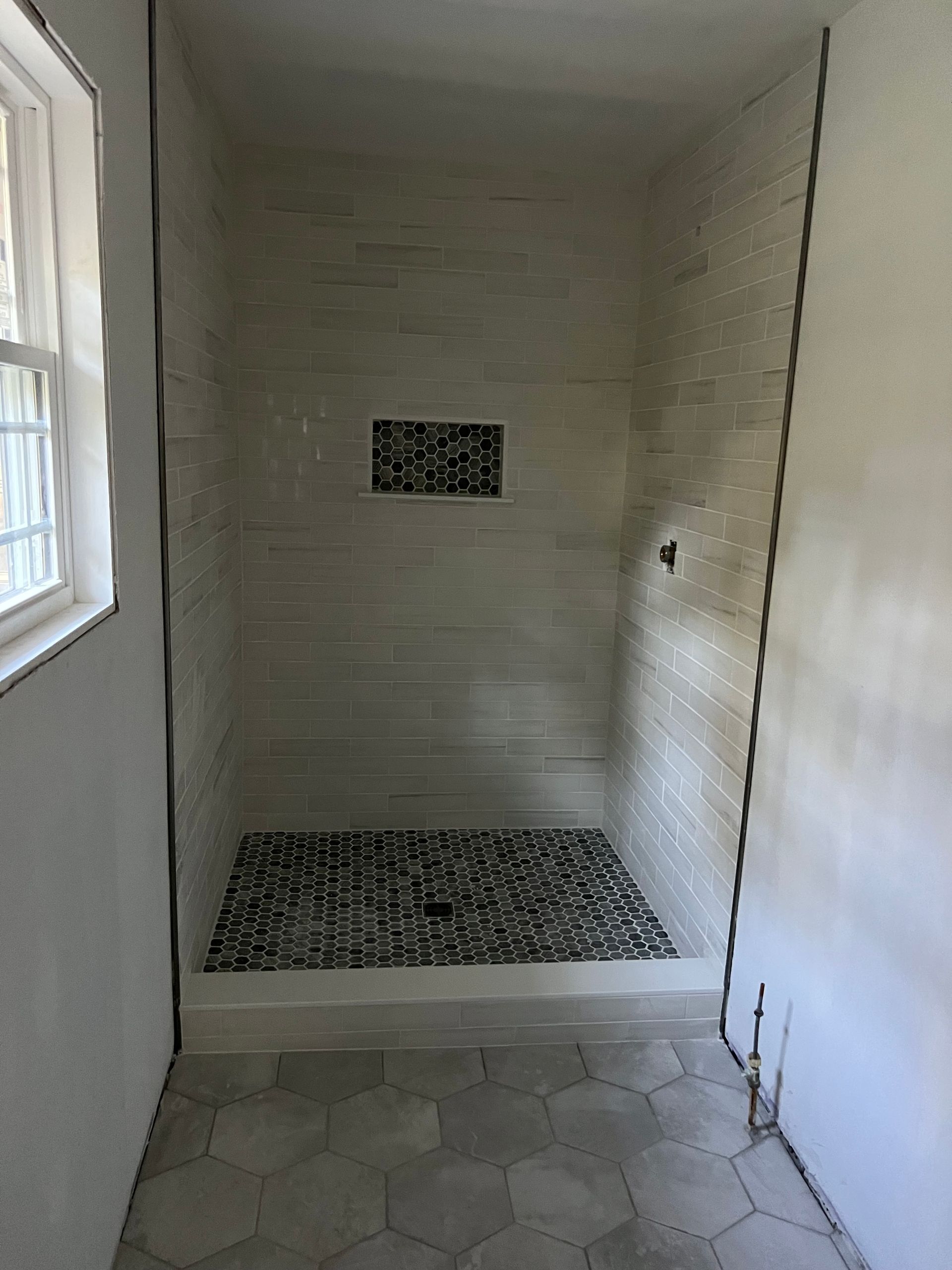 Narrow white shower stall with tiled floor, wall niche, and small window on the left.