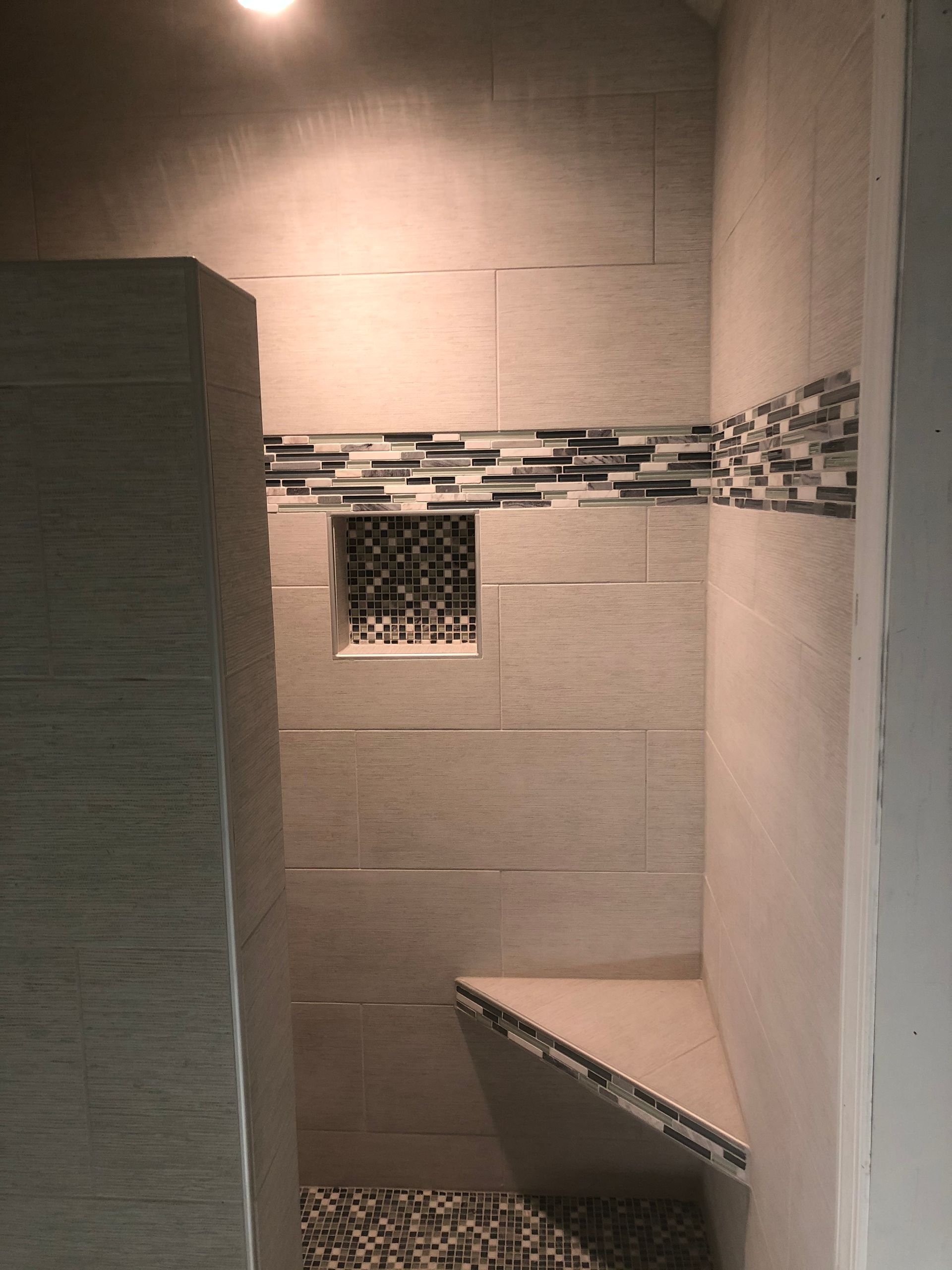 Narrow tiled shower stall with mosaic accent strip, small window, and corner bench seat.