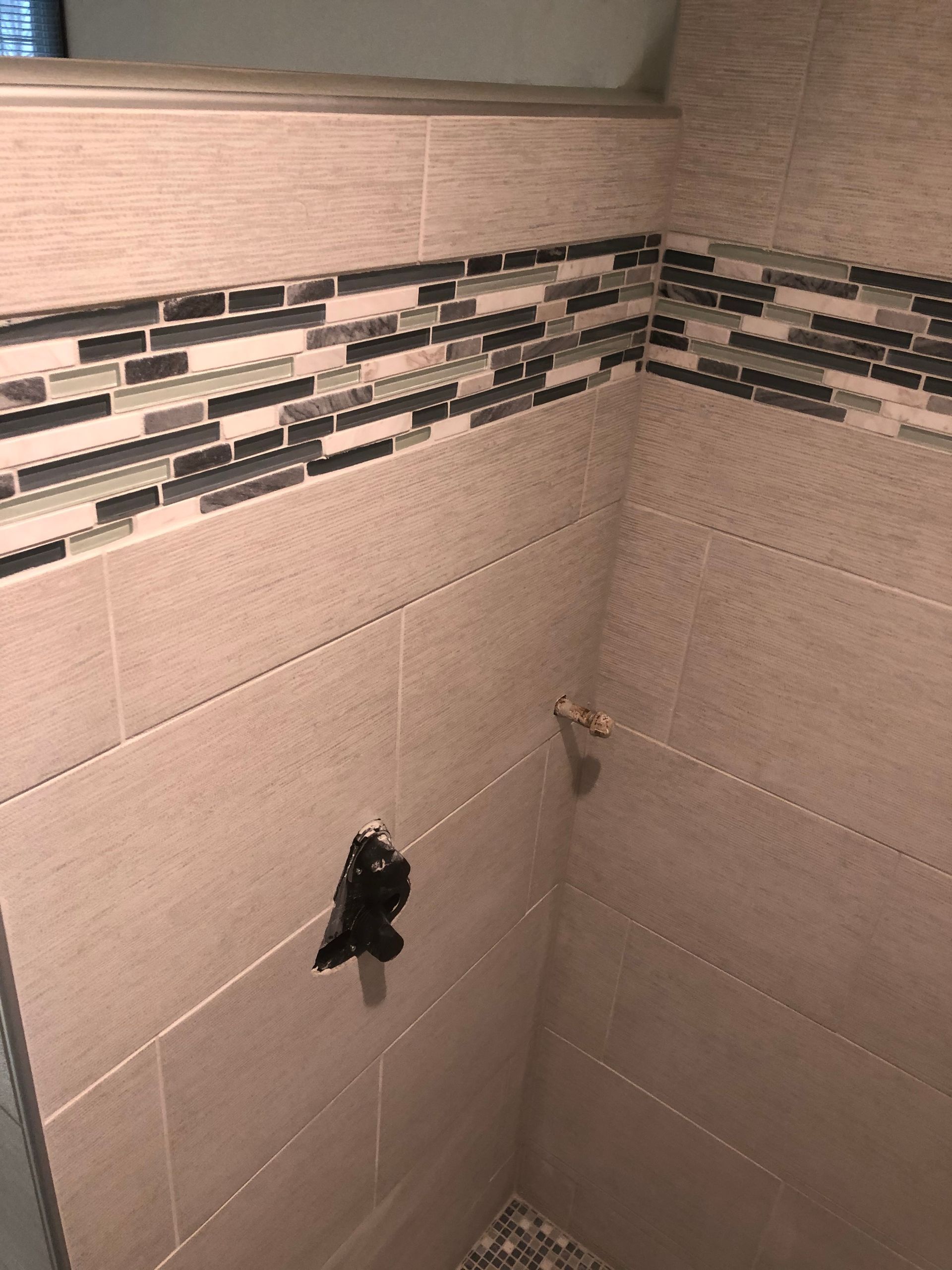 Tiled bathroom wall with decorative border and exposed plumbing fixtures and holes.