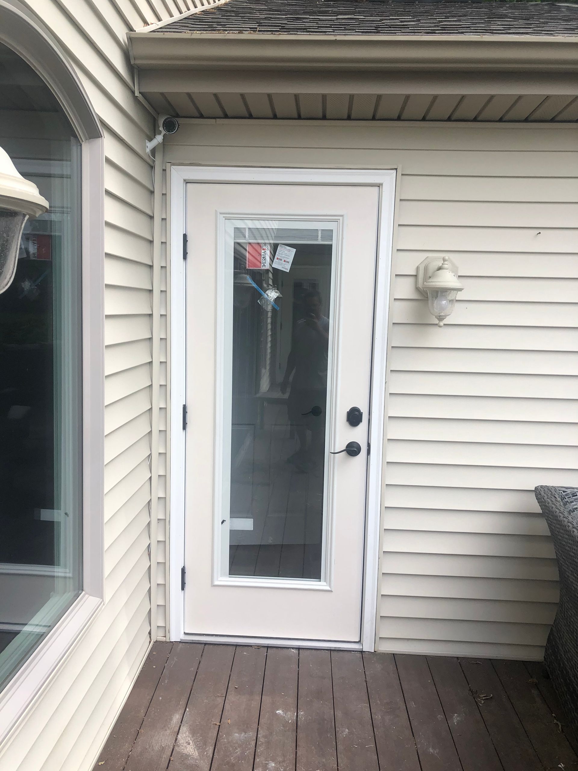 White exterior door with glass panel on a wooden deck beside beige siding, next to a window and wall light.