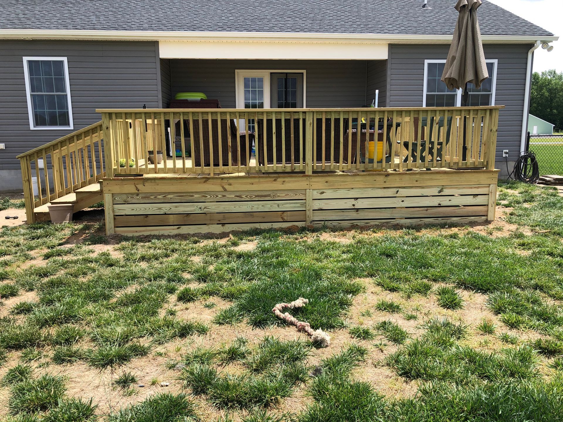 Backyard deck with wooden railing and stairs, behind a gray house with a patchy grass yard.