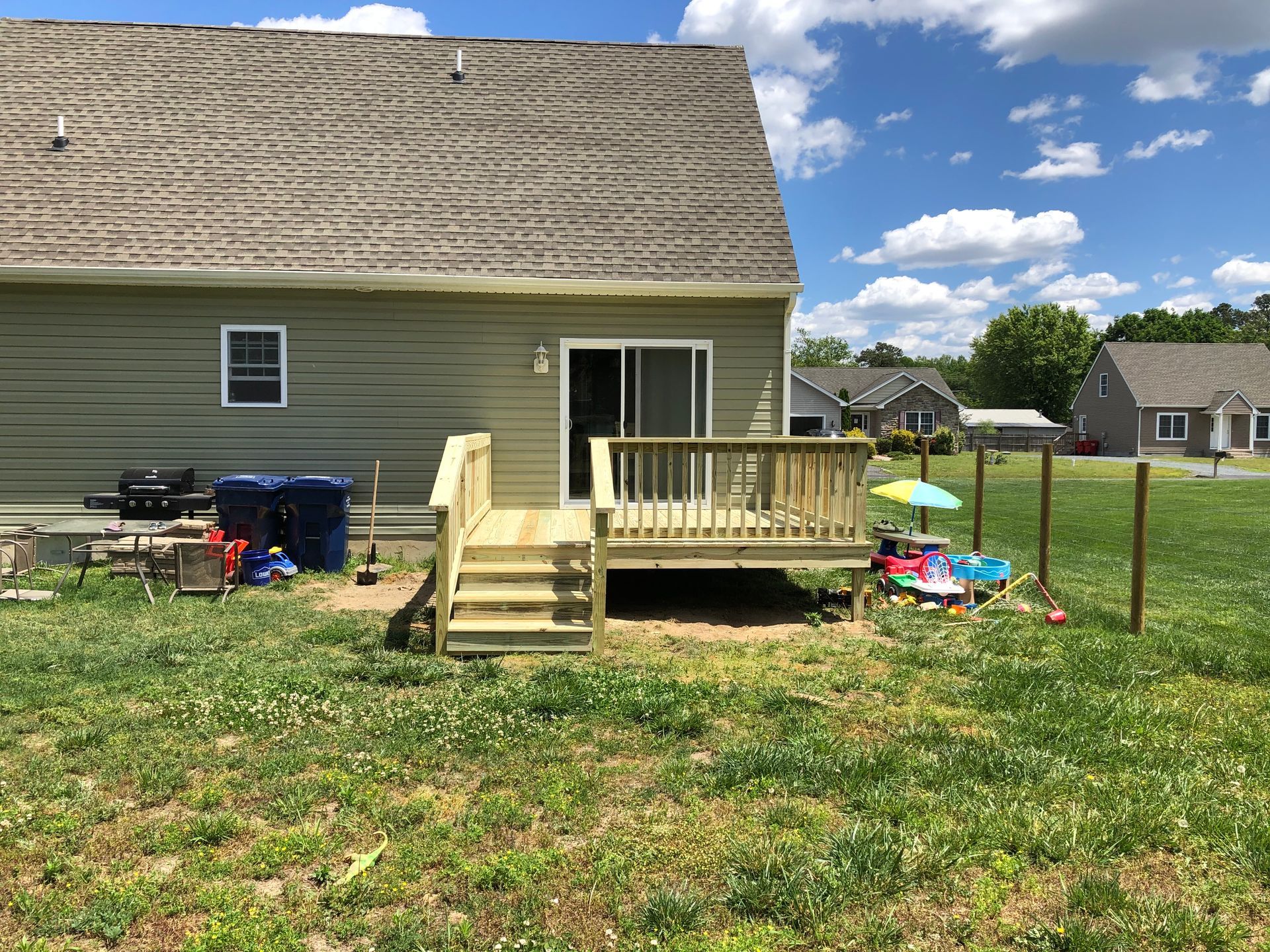 Backyard with a house deck, steps, and lawn; scattered outdoor items and a fenced yard under a partly cloudy sky