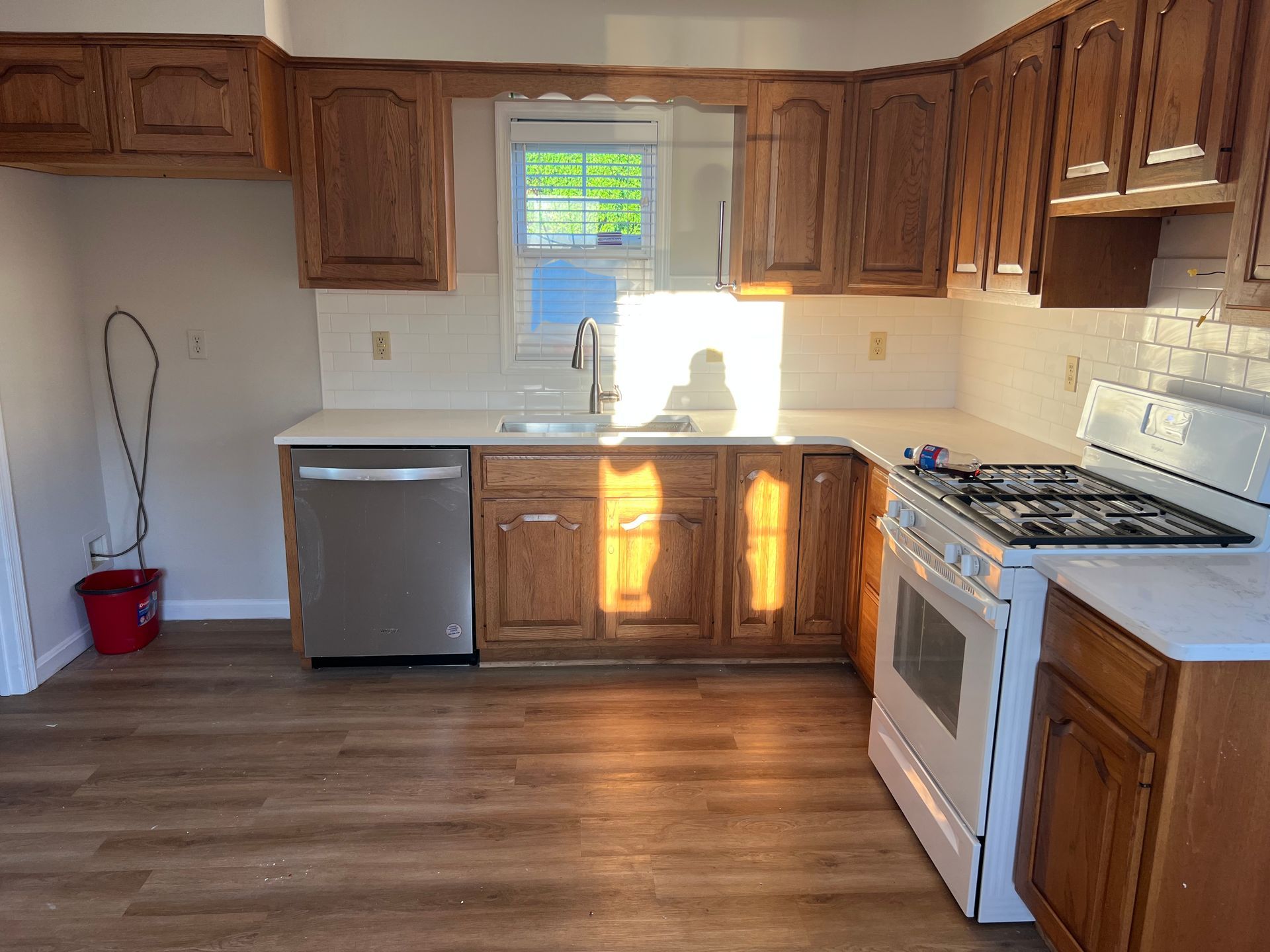 Empty kitchen with wooden cabinets, white stove, stainless dishwasher, and sunlit window over the sink.