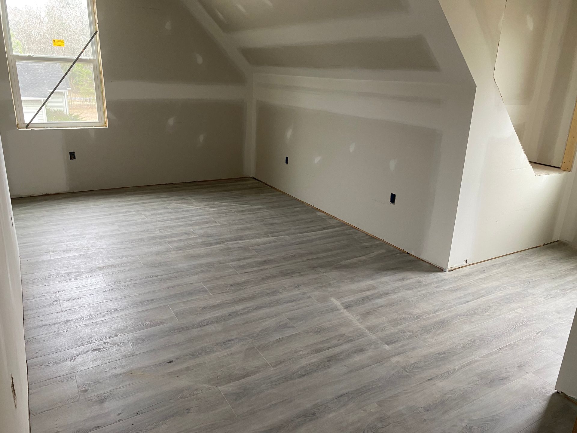 Empty attic room with sloped white walls, gray wood floor, and windows letting in daylight