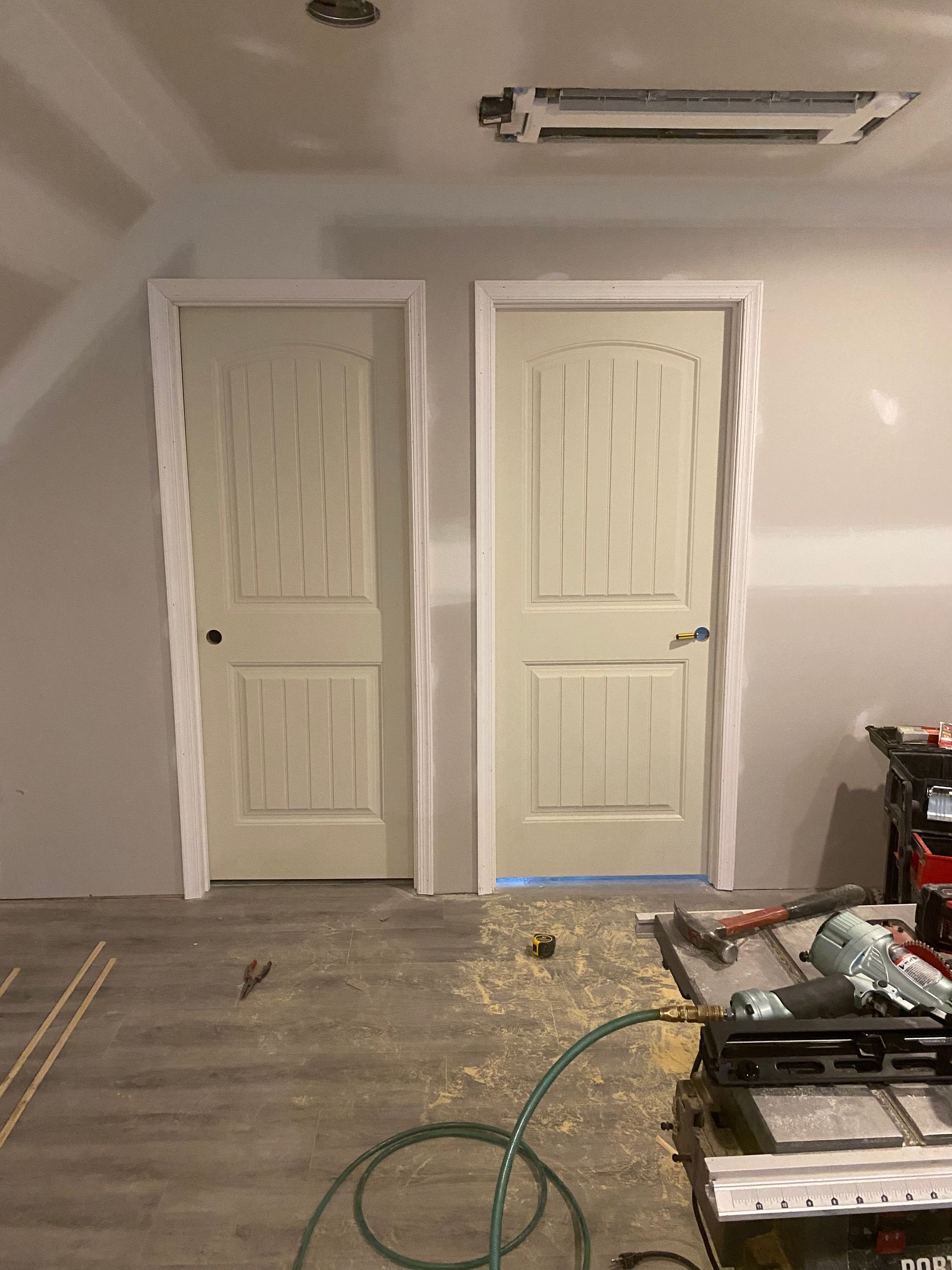 Interior room with two white doors in a beige wall, exposed ceiling fixture, and construction tools on the floor.