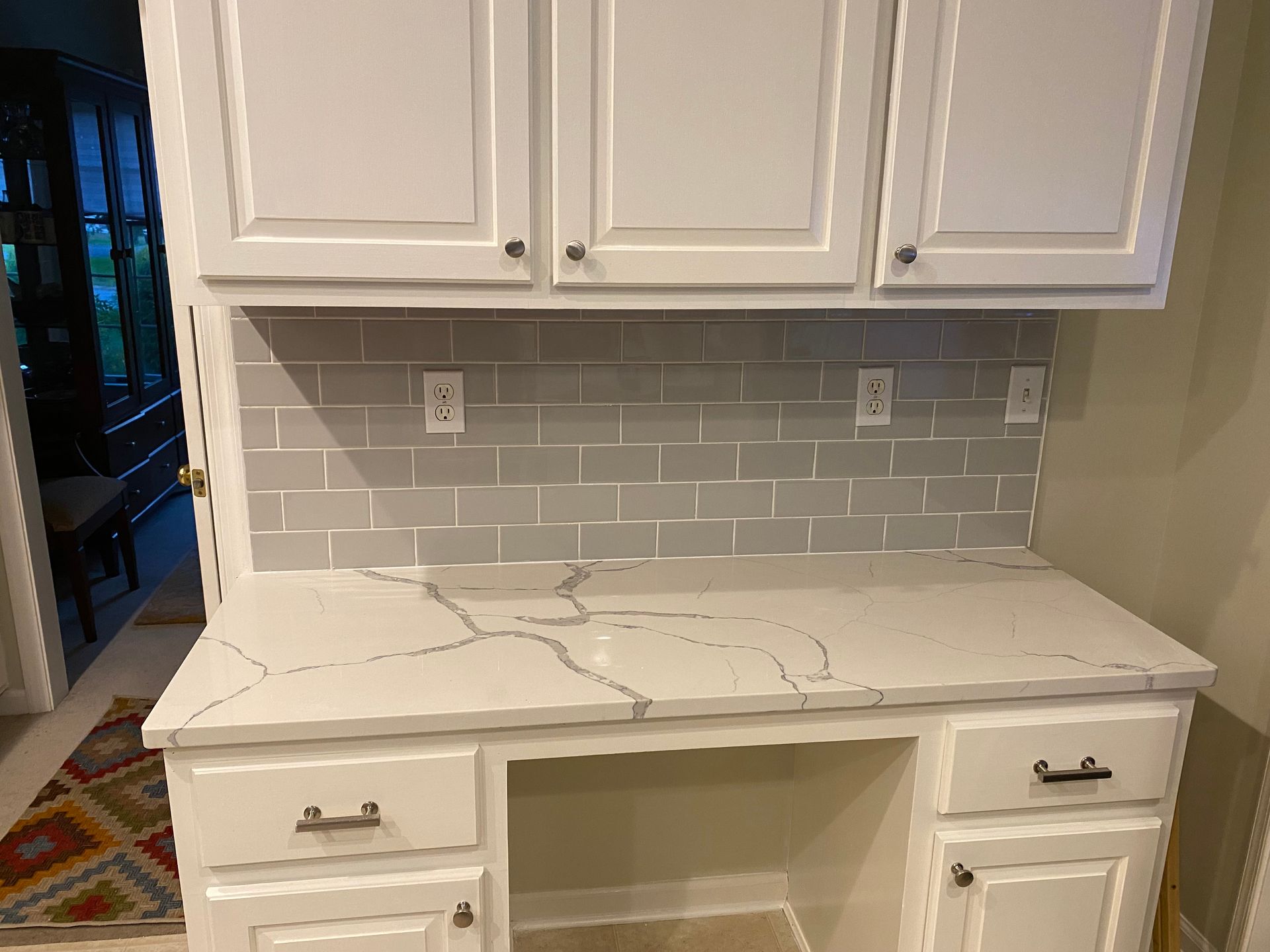 White built-in desk with marble-look top and cabinets against a gray subway-tile backsplash.