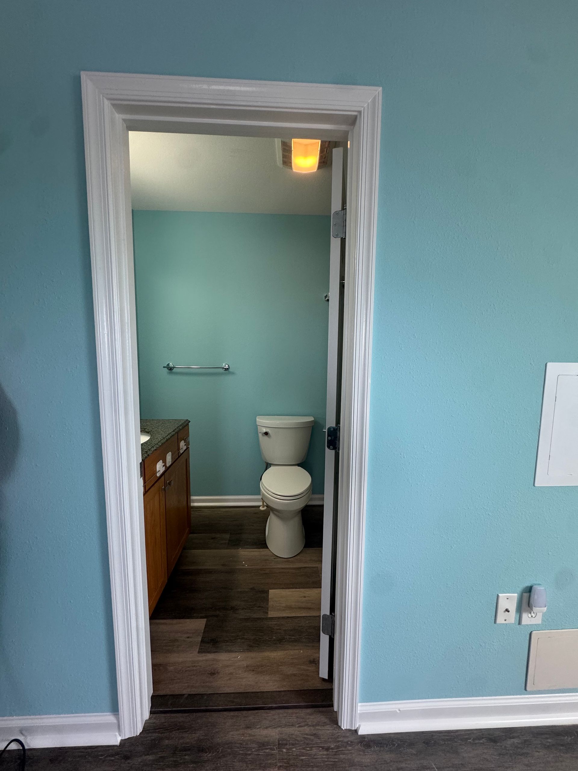 View into a small bathroom with aqua walls, wood vanity, and a toilet under a lit ceiling fixture.