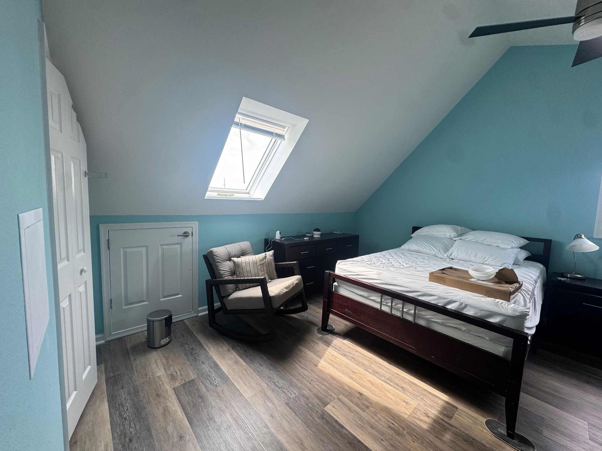 Small attic bedroom with blue walls, skylight, bed, chair, and dresser/desk area