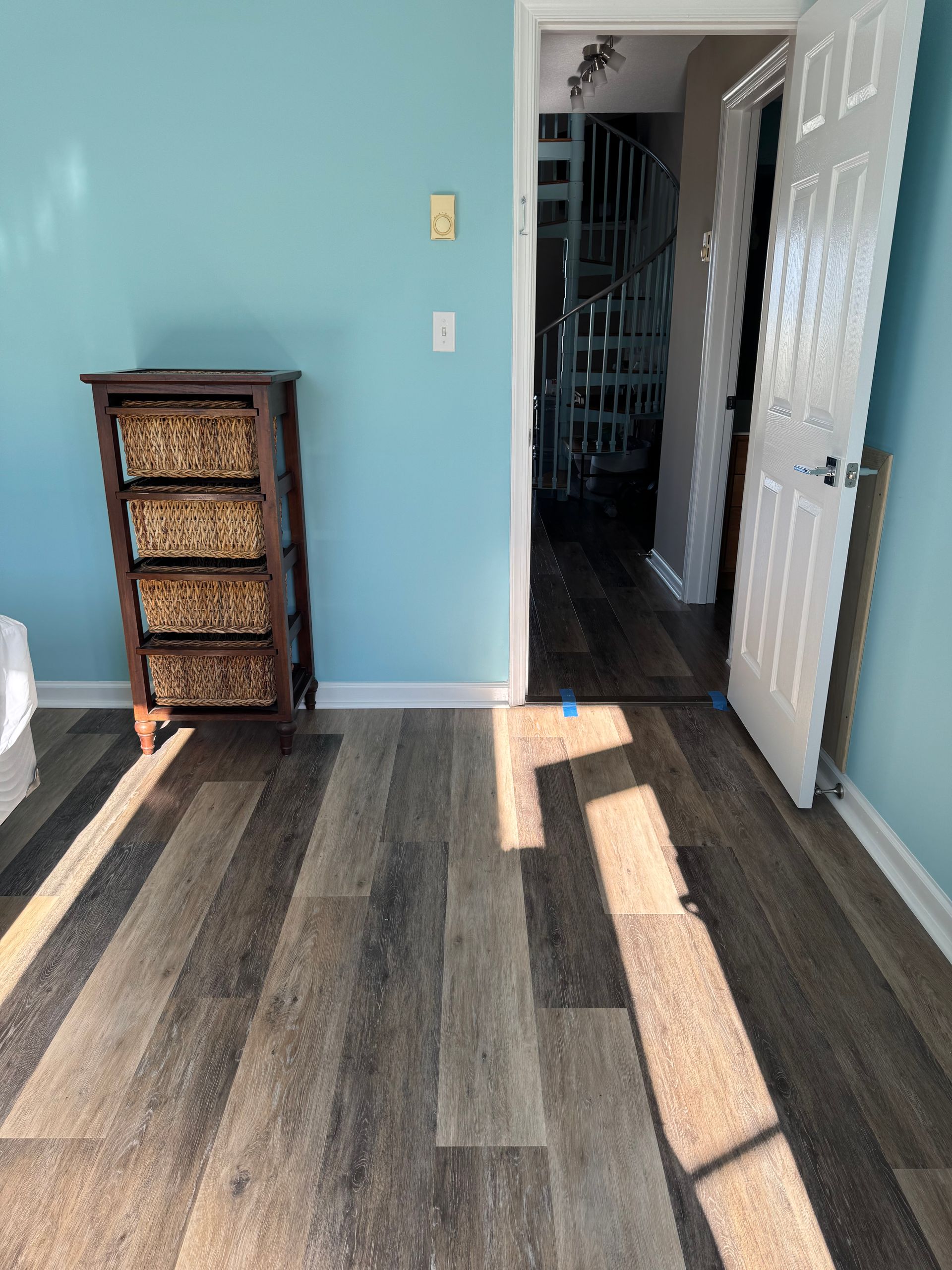 Empty room with gray wood floors, blue wall, wicker drawer unit, and sunlight near an open white door