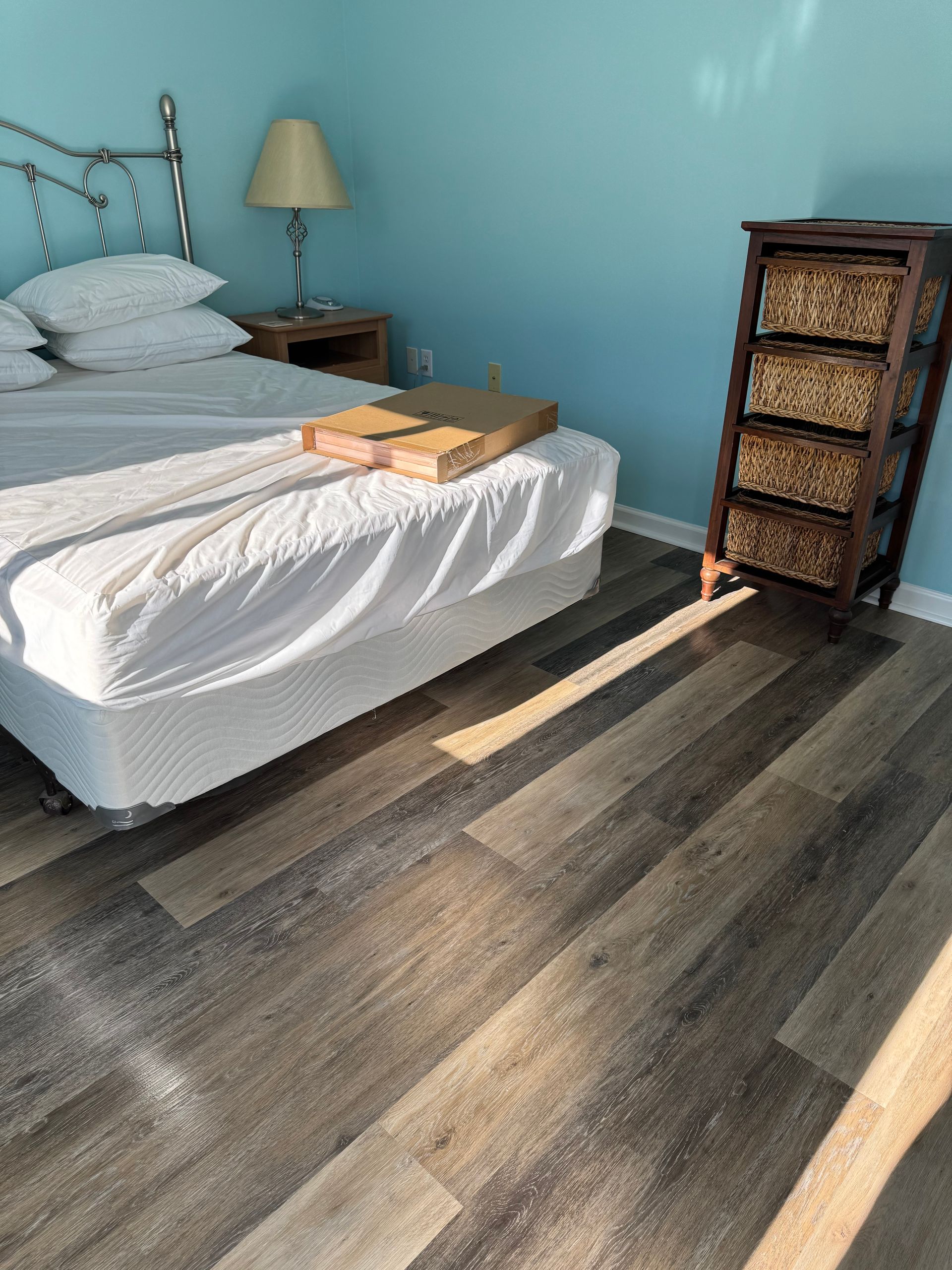 Bedroom with white bed, bedside lamp, woven drawer chest, and sunlight on dark wood floor.