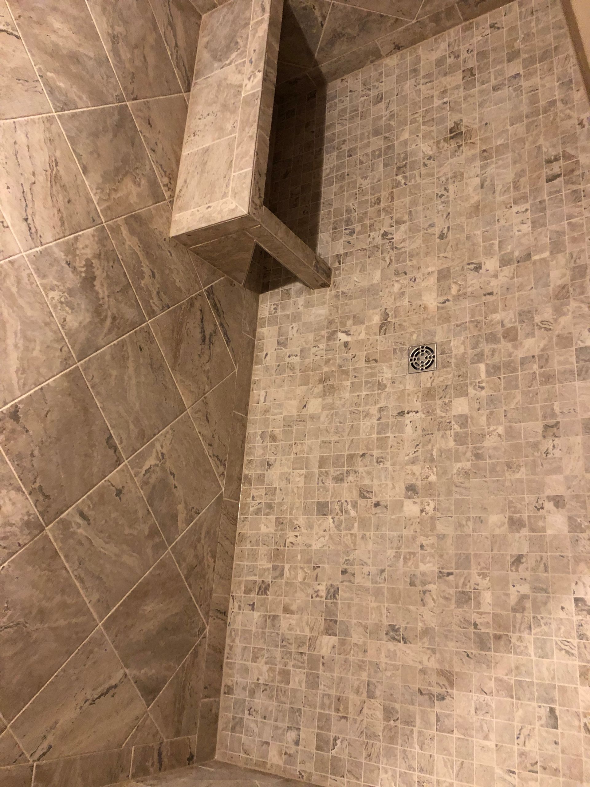 Tan tiled shower stall with a built-in shelf and a drain on the floor.