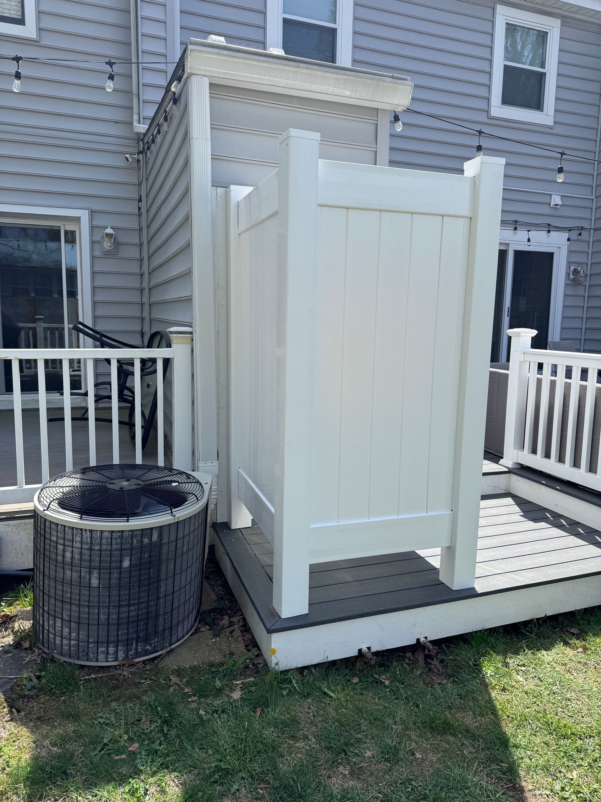 White outdoor shower stall beside a gray house on a small deck, next to a planter and white railing.