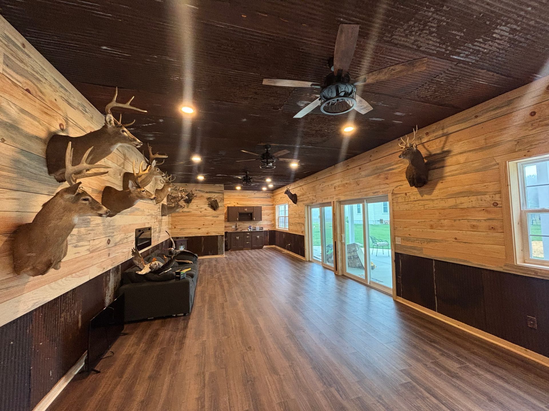 Wide rustic game room with mounted deer heads, wood-paneled walls, ceiling fans, and large windows.