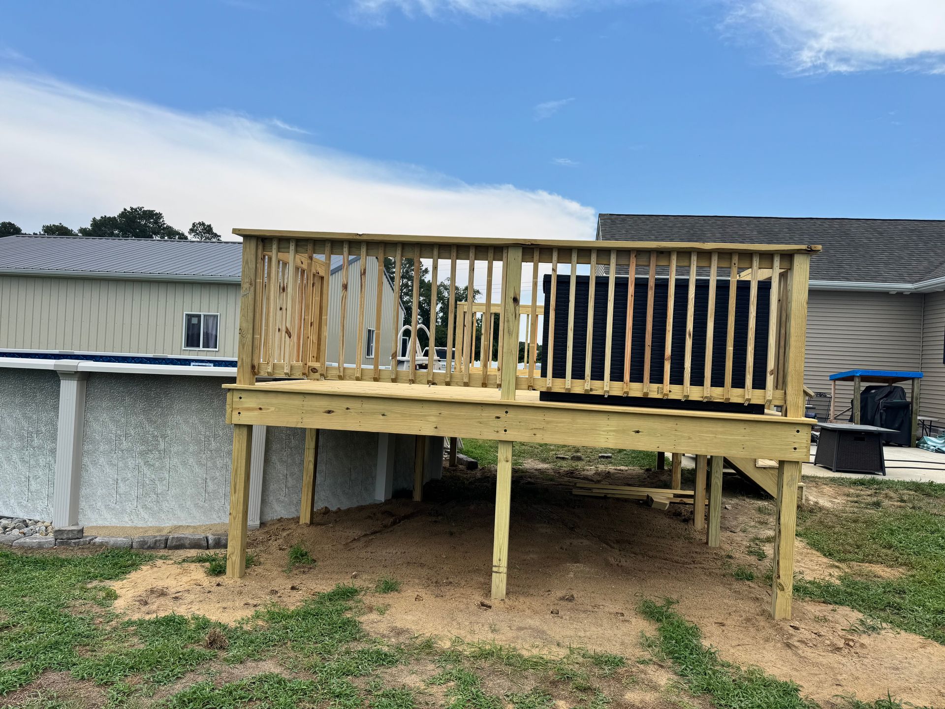 Wooden raised deck under construction beside a mobile home on a dirt yard.