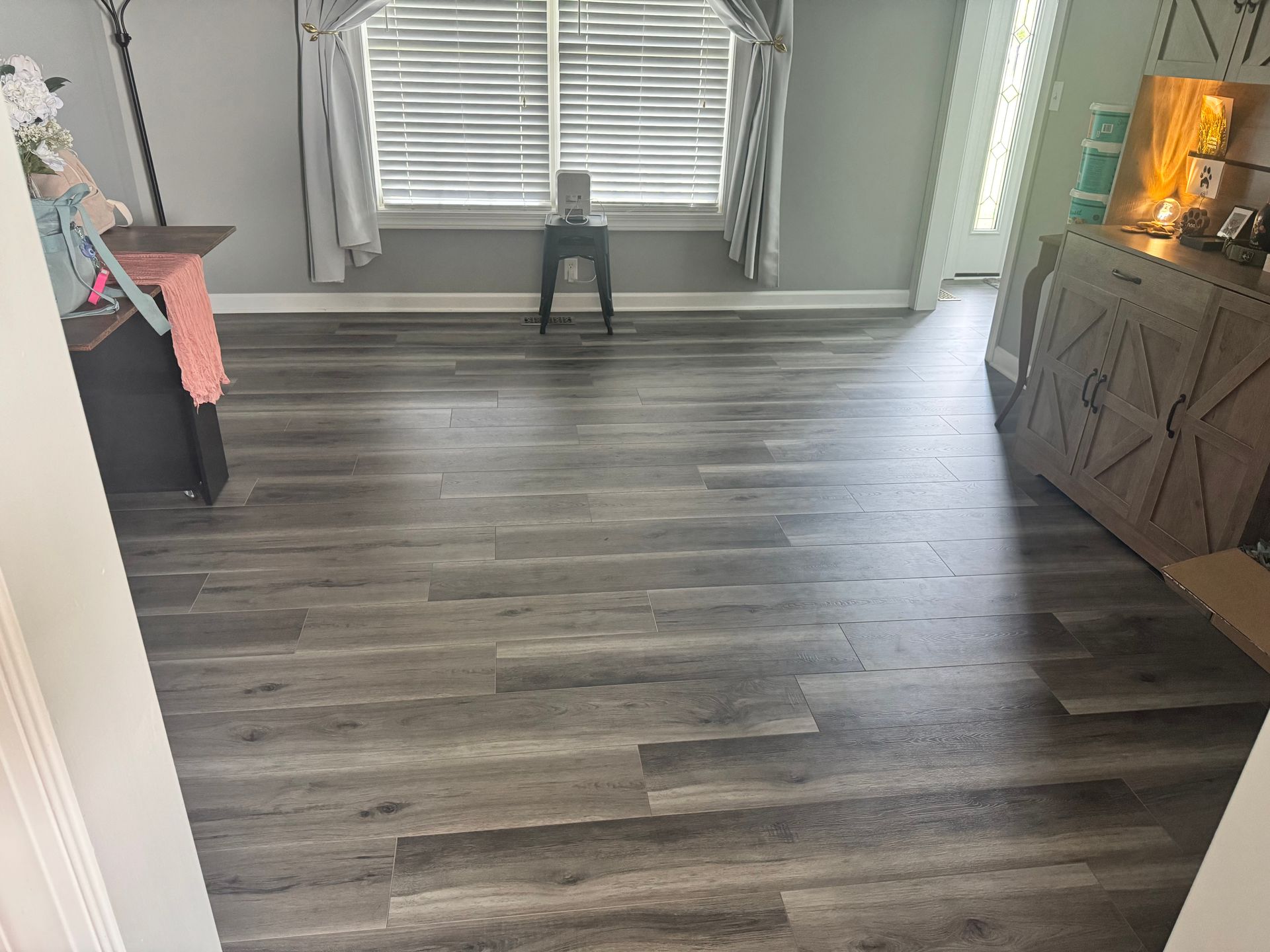Empty living room with gray wood flooring, large window blinds, and a cabinet on the right.