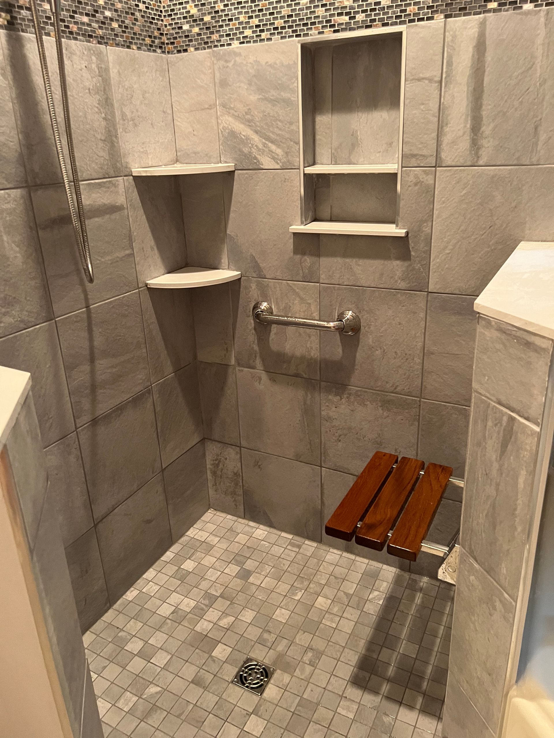 Small tiled shower stall with built-in shelves, grab bars, and a wooden bench seat.