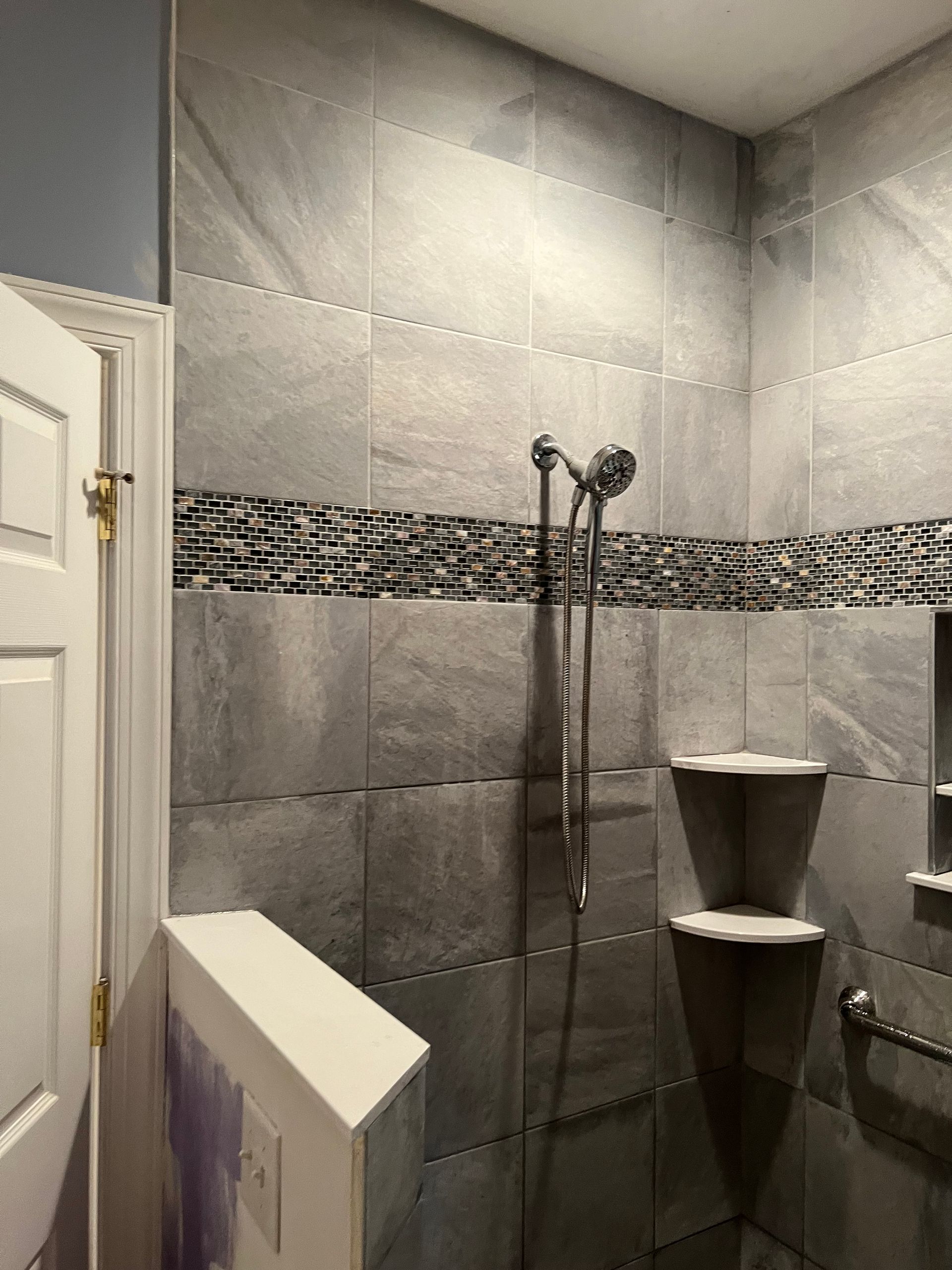 Tiled bathroom shower with glass partition, gray walls, and corner corner shelves.