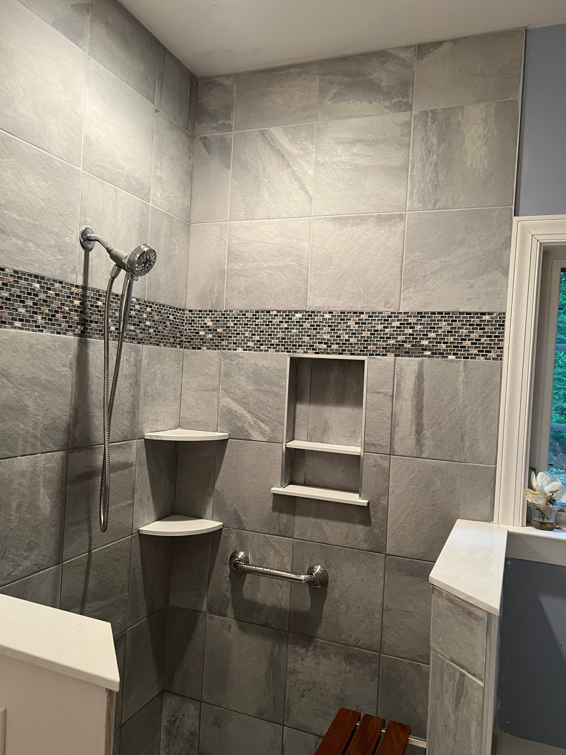 Gray tiled bathroom shower with corner shelf, mosaic border, and handheld showerhead