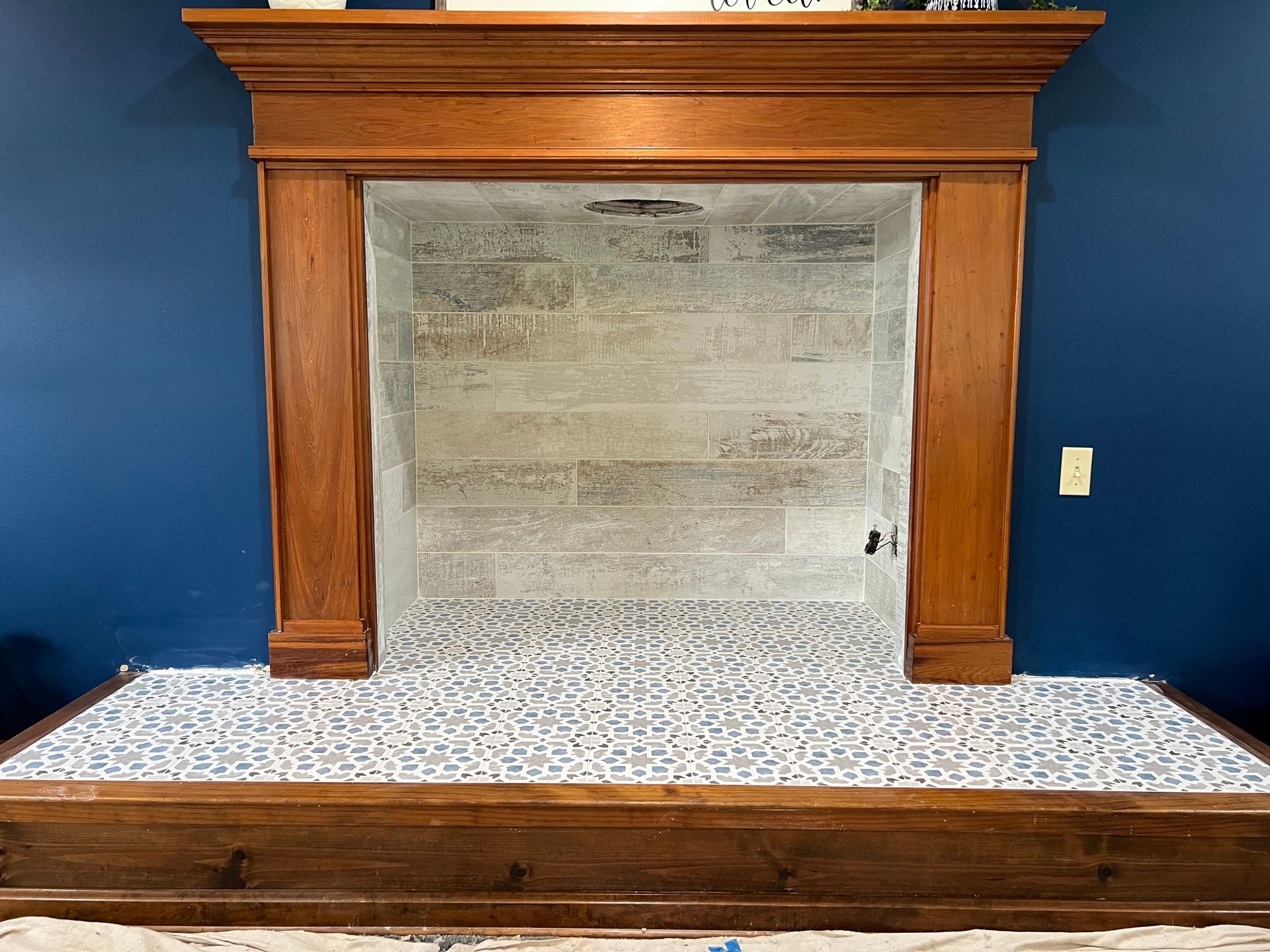 Wooden fireplace mantel with a gray stone firebox and blue wall backdrop, over a patterned tile hearth.