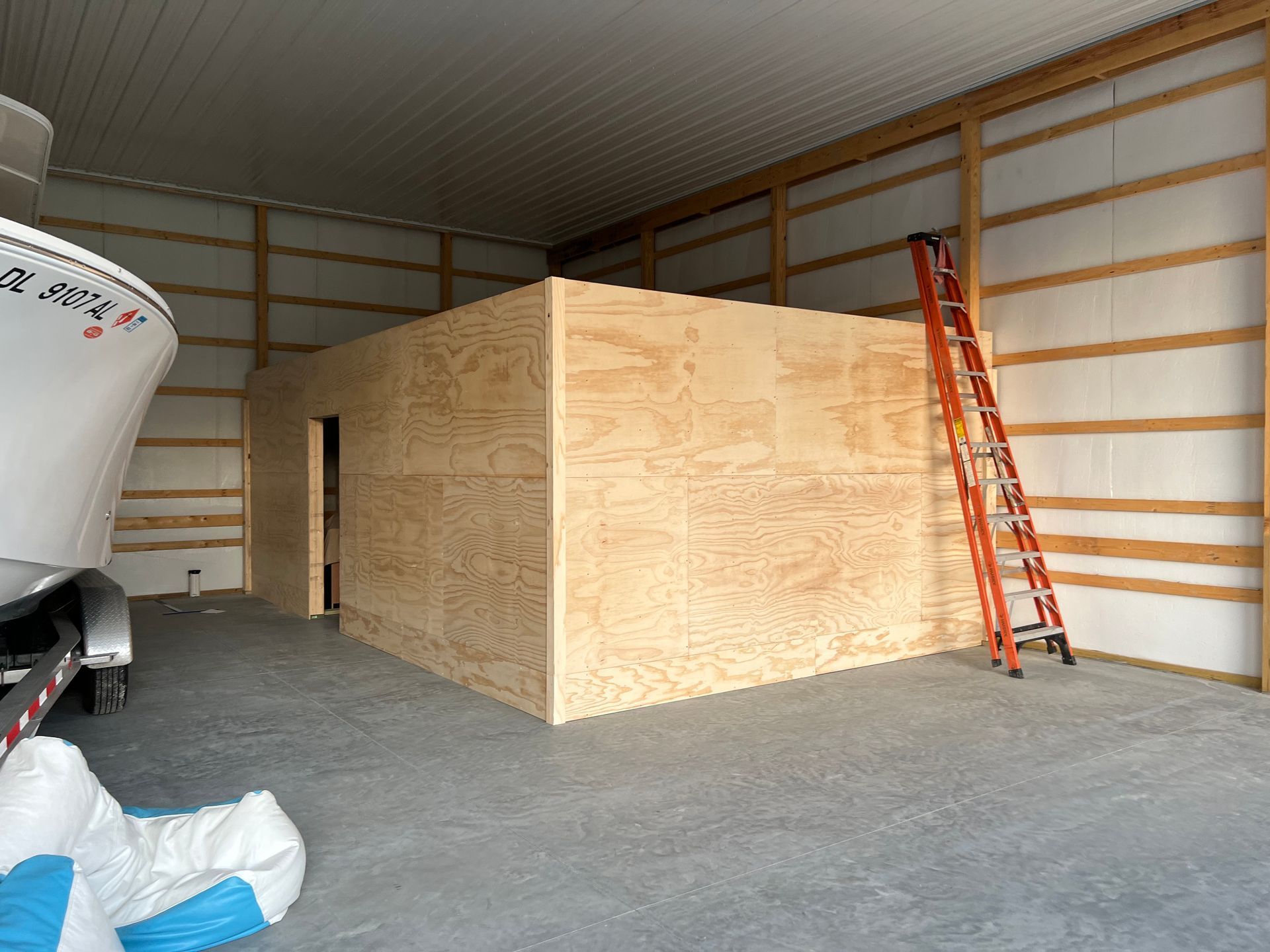 Plywood-walled room inside a warehouse, with a ladder leaning on the right and a boat at left.