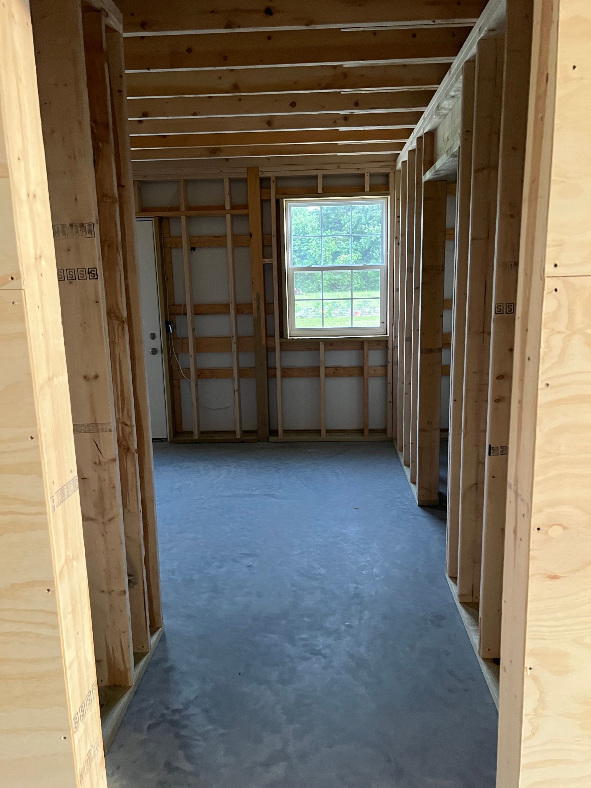 Framed room interior with exposed wood studs, concrete floor, and a window at the far end.