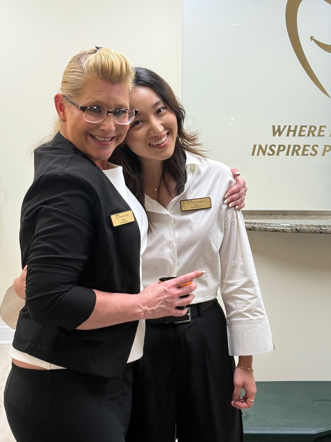 Two women in business attire pose, smiling, embracing. One has blonde hair, glasses, the other has dark hair.