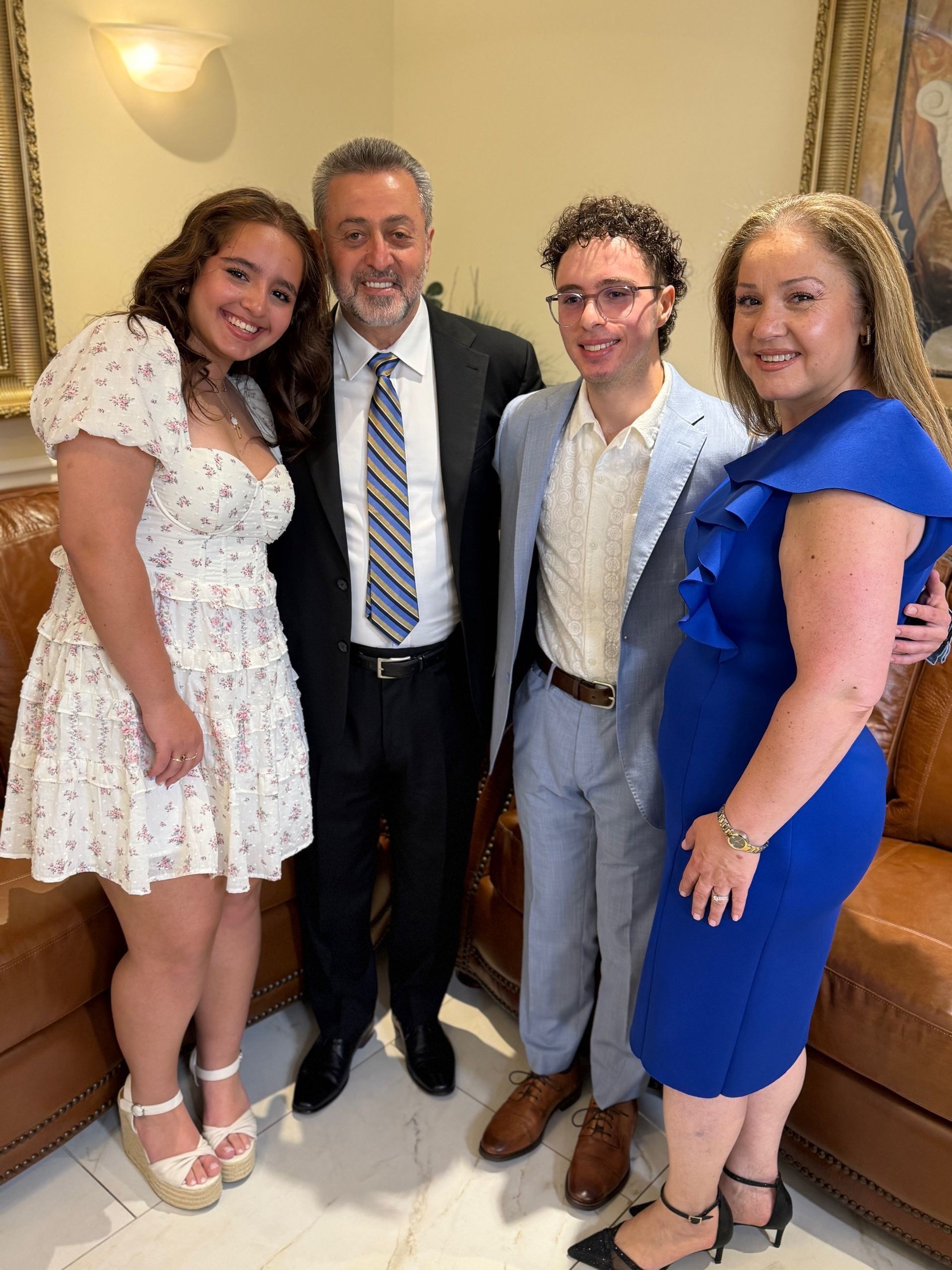 Family of four poses indoors. Woman in blue dress, man in suit, girl in floral dress, boy in light blue suit.