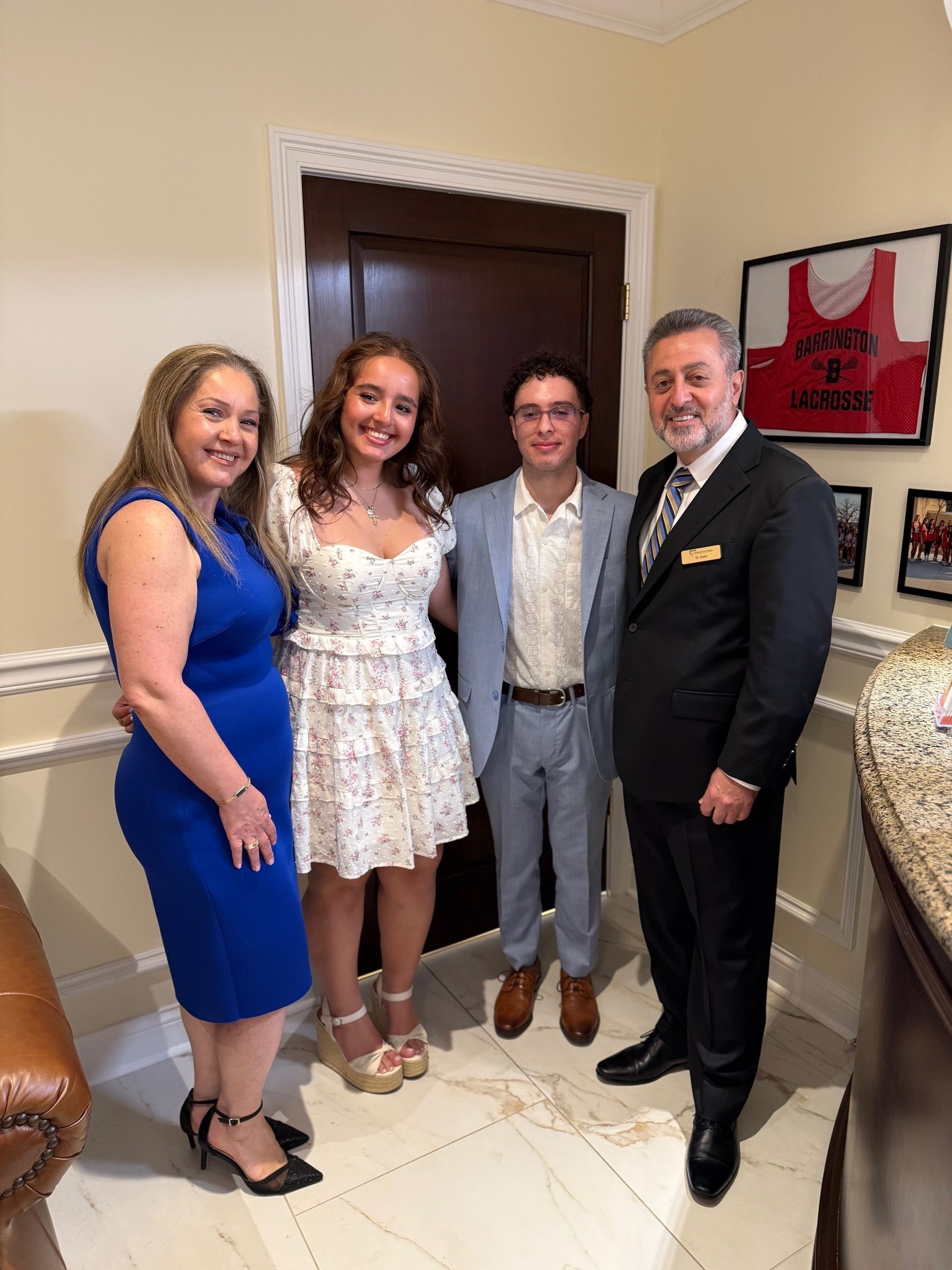 Family poses for a photo near a doorway. Woman in blue dress, teen in floral dress, teen in suit, man in suit.