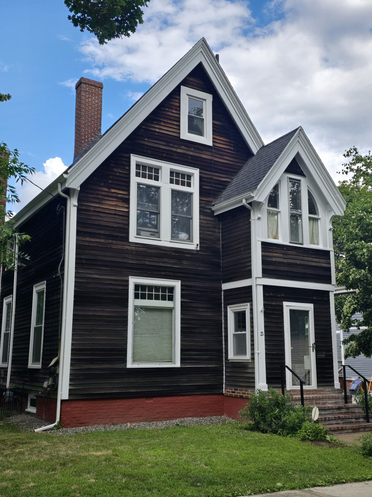 Brown two-story house with white trim, dark roof, brick chimney, and red foundation. Green lawn and steps lead to door.