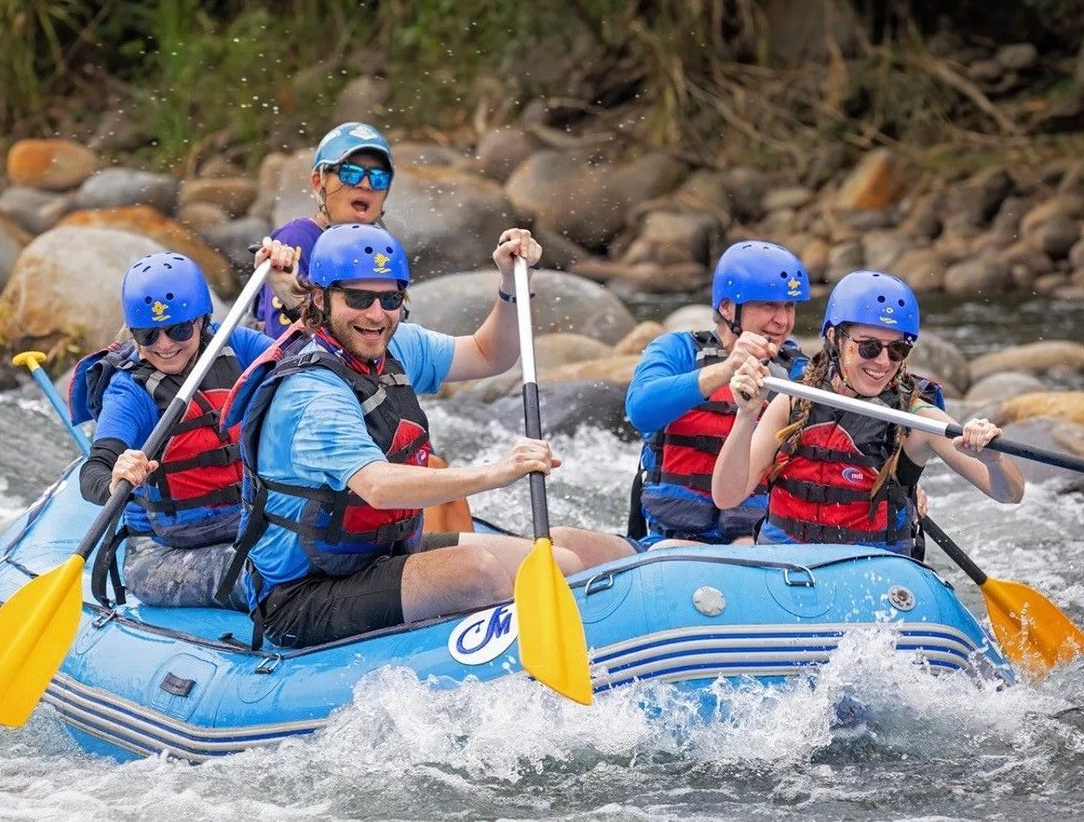 People rafting down a river, paddling and smiling. Blue raft, helmets, life vests.