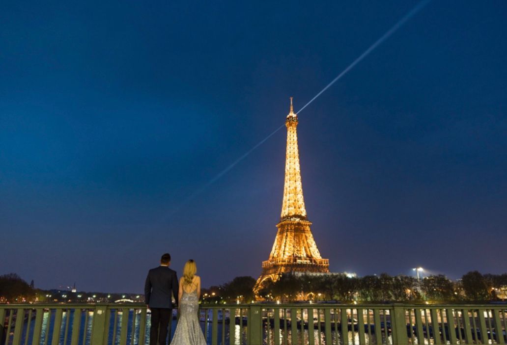 Couple admiring the Eiffel Tower at night; bride in white gown, groom in suit; Paris skyline with lights.