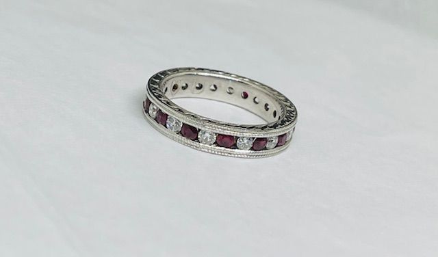 Silver eternity ring with alternating red gemstones and clear stones, resting on a white surface.