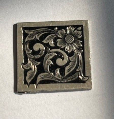 Square silver metal tile with floral engraved pattern on a black background.