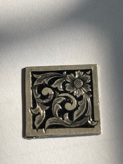 Silver square with floral and scroll engraving, black background.