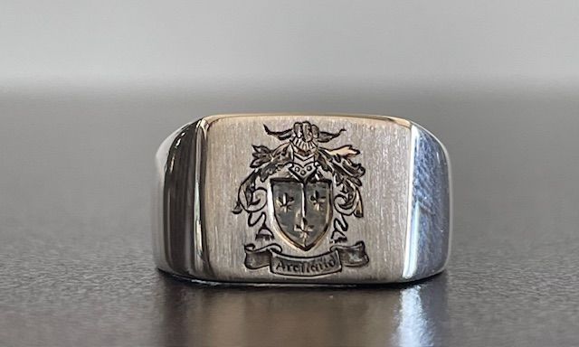 Silver signet ring with engraved family crest, resting on a reflective surface.