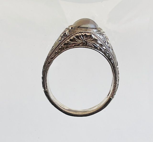 Detailed silver ring with a large, round, light-colored gemstone, engraved floral design around the band.