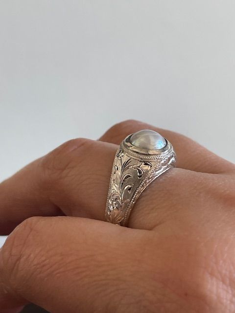Close-up of a hand wearing a silver ring with an engraved band and a large, round, pearly gem.