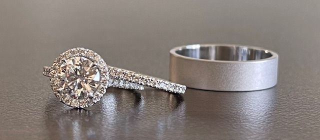 Two silver rings, one with a large diamond and a halo of smaller diamonds, the other a plain band, sit on a surface.