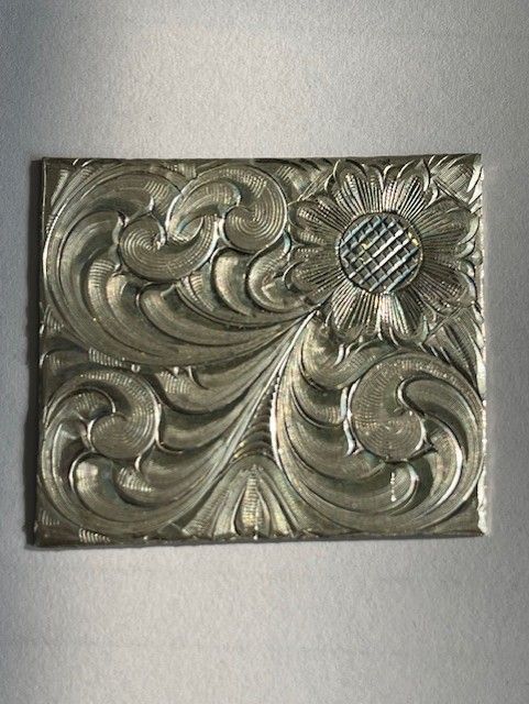 Silver metal engraved with floral scrollwork and a textured flower in a rectangle.