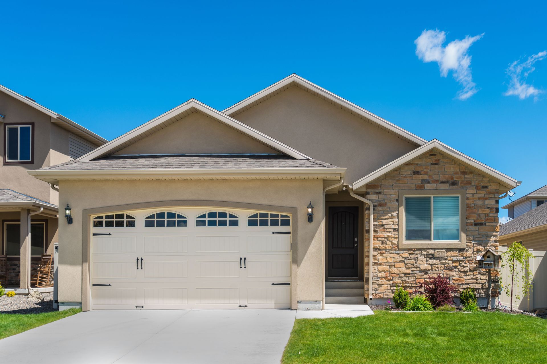 Tan house with a stone facade, beige garage door, and green lawn under a blue sky.