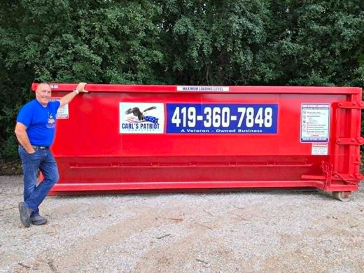 Man standing beside a red dumpster with a large 419-360-7848 sign, outdoors by trees
