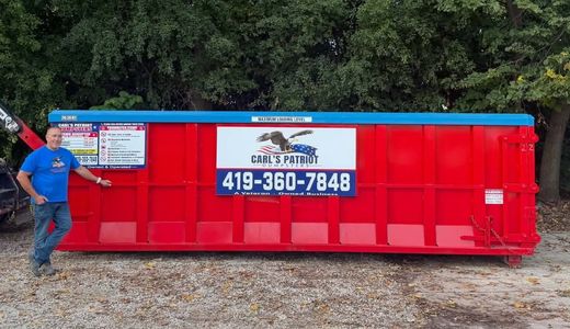 Red dumpster with company sign and phone number; man in blue shirt stands beside it outdoors.
