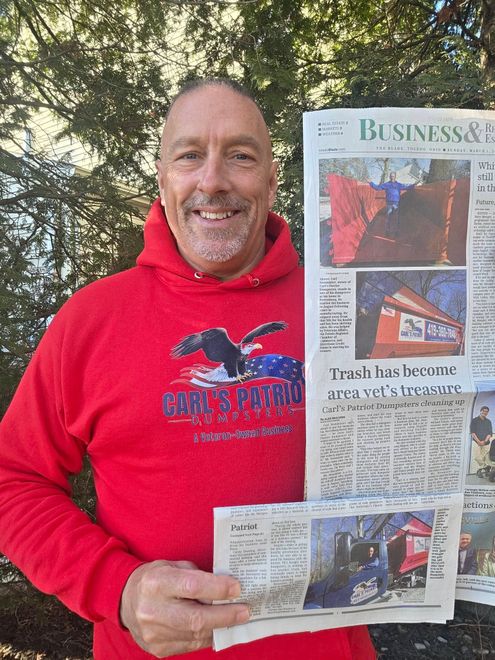 Man in red hoodie holding a newspaper outdoors, smiling at the camera.