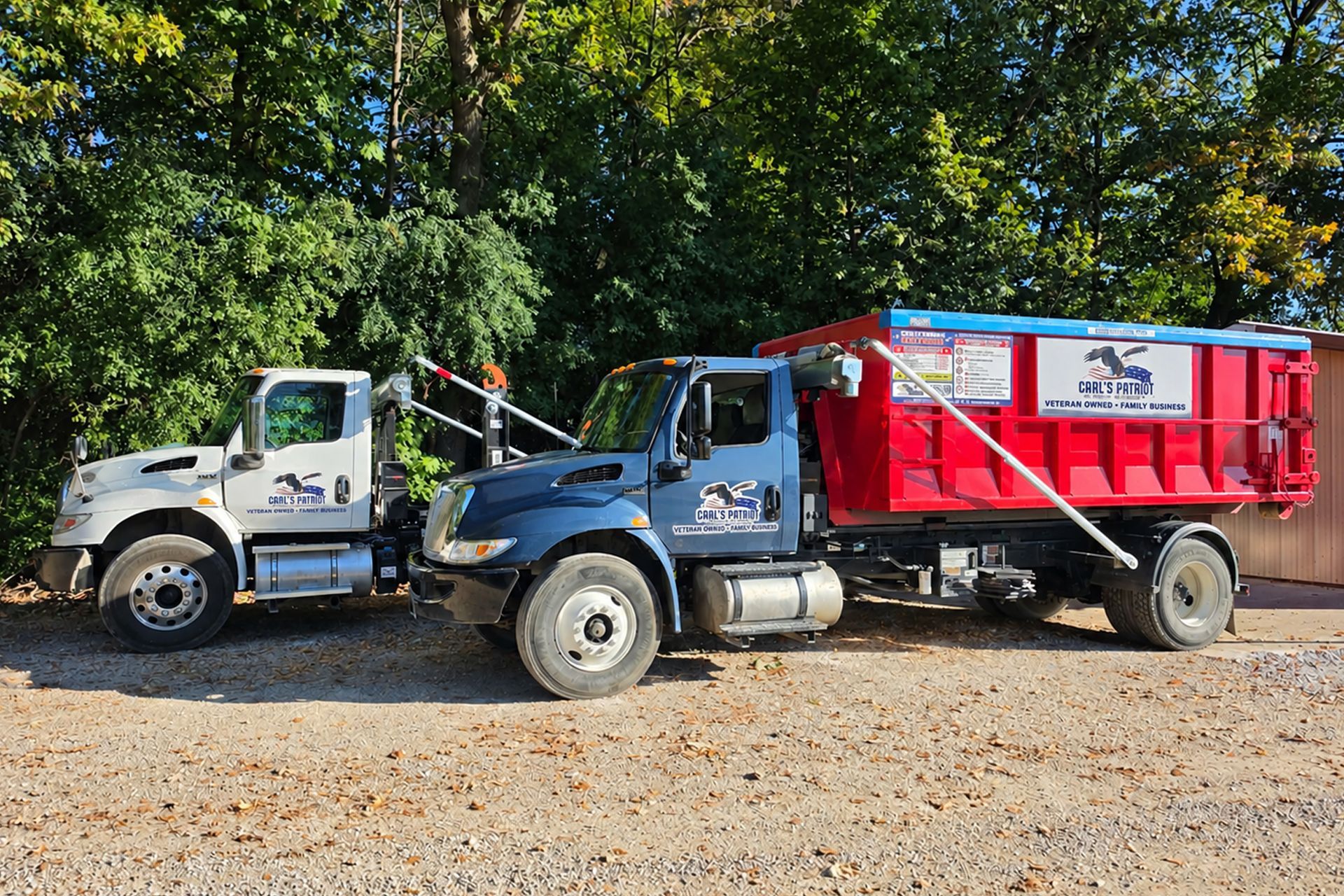 Two dump trucks, one white and one blue with a red bed, parked on gravel under trees.