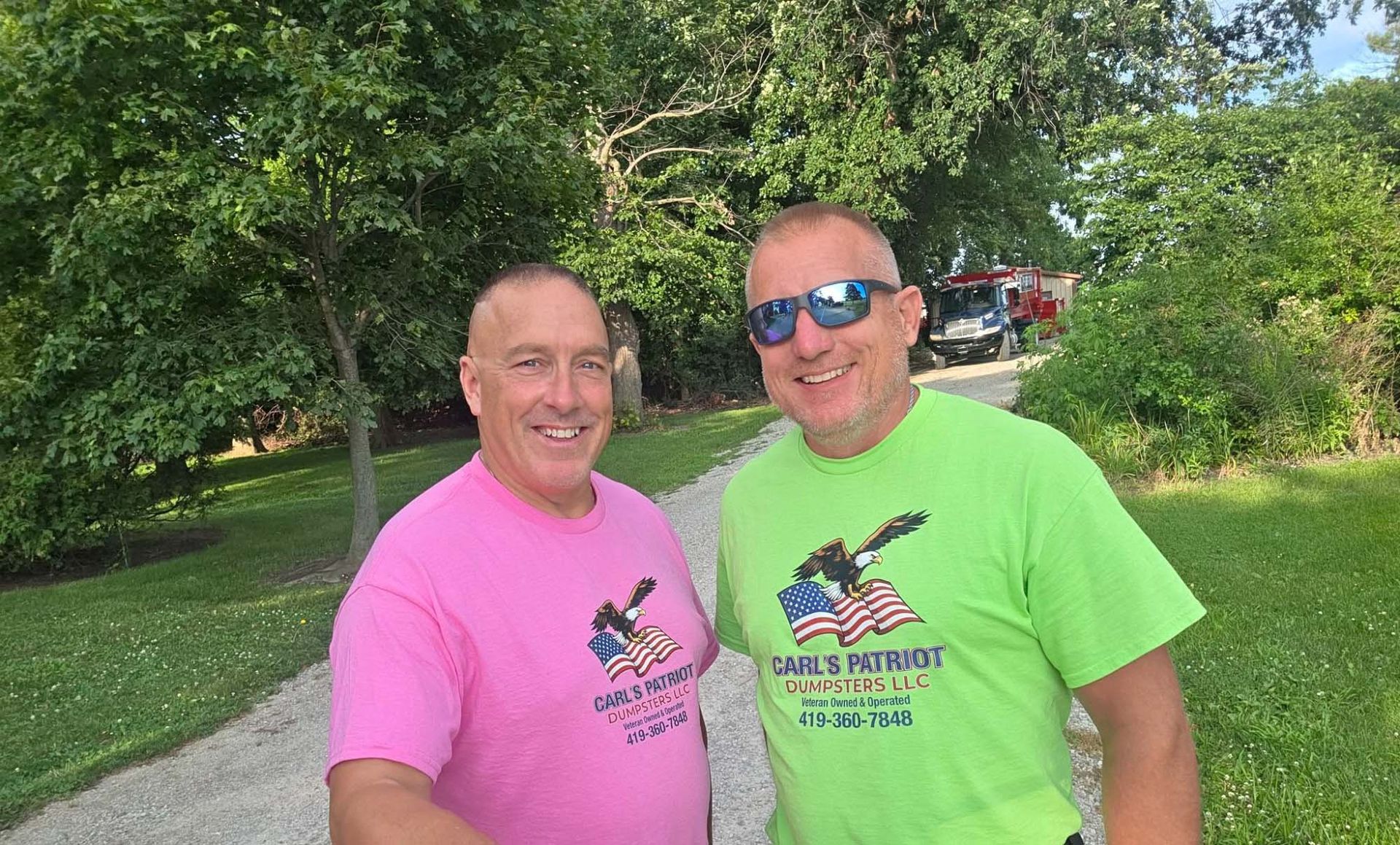 Two smiling men in pink and green shirts standing outdoors on a sunny lawn.