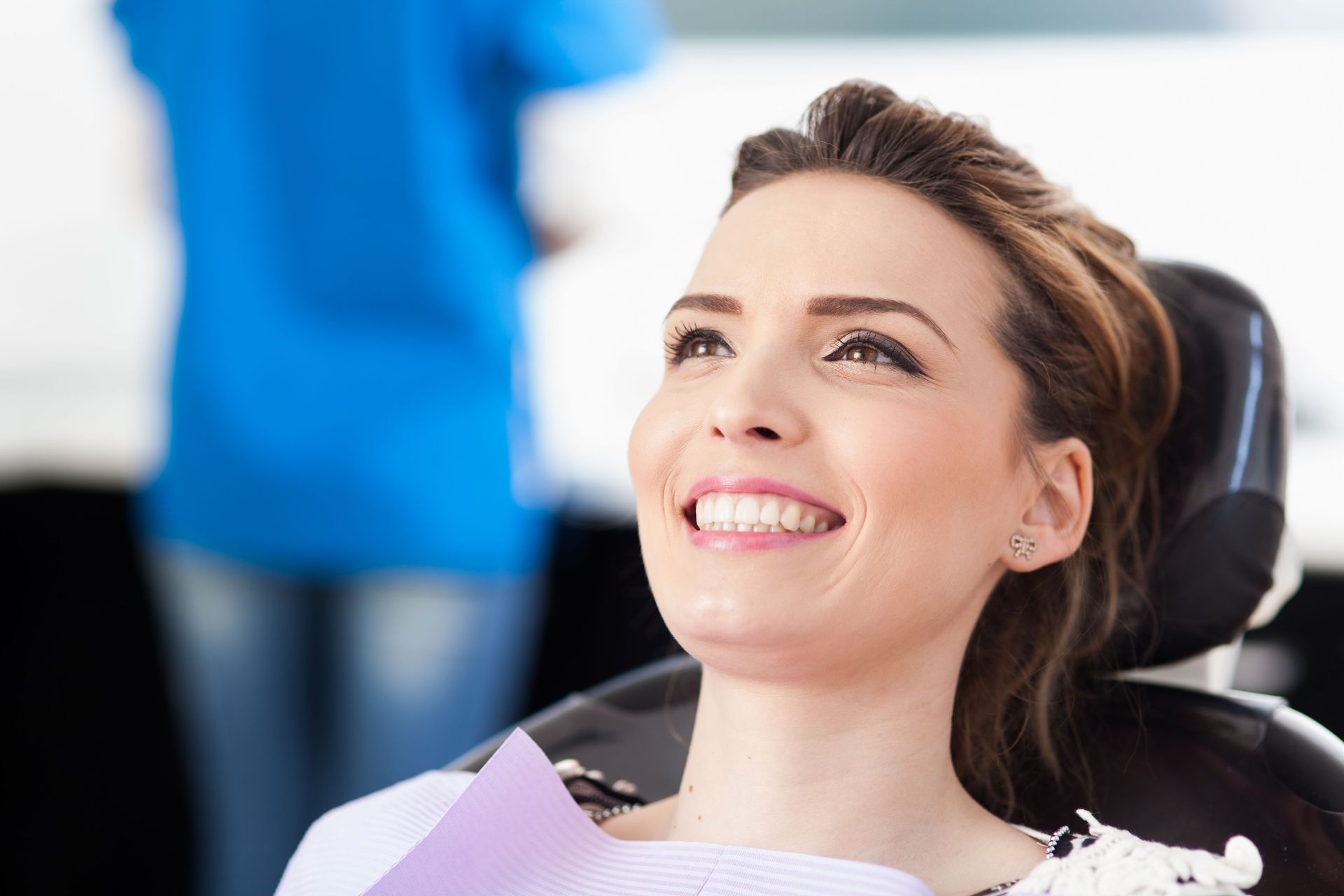 Smiling patient in a dental chair with a blurred dentist in blue scrubs behind her